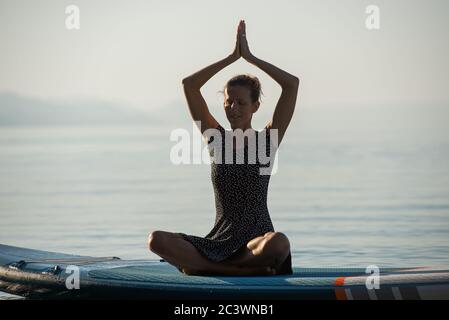 Jeune femme sereine avec un sourire sur son visage assis en position lotus avec ses paumes jointes au-dessus de sa tête comme elle pratique la méditation et le yoga outsid Banque D'Images