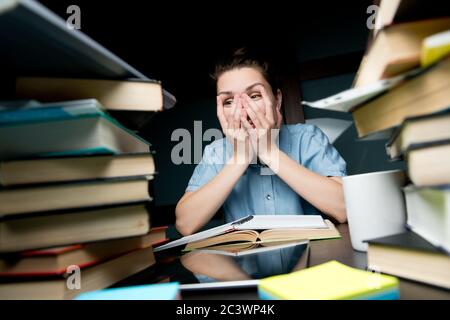 La jeune fille étudiante regarde avec lassitude à travers ses doigts sur le montagne de livres Banque D'Images