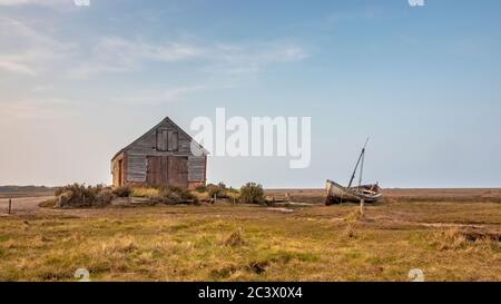 L'ancienne grange à charbon du port de Thornham, sur la côte nord de Norfolk, avec un vieux bateau de pêche abandonné et abandonné, qui se trouve d'un côté au soleil du printemps Banque D'Images