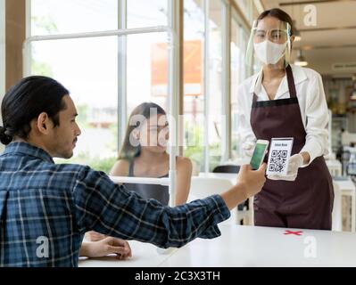 Un client asiatique lit le menu en ligne du code QR depuis une serveuse avec masque et masque facial. Le client s'est assis sur une table de danse sociale pour un nouveau style de vie normal Banque D'Images