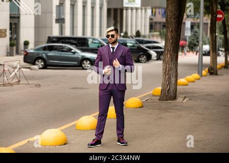 homme en lunettes et costume trois pièces violet de luxe, noeud papillon posant dans la rue Banque D'Images