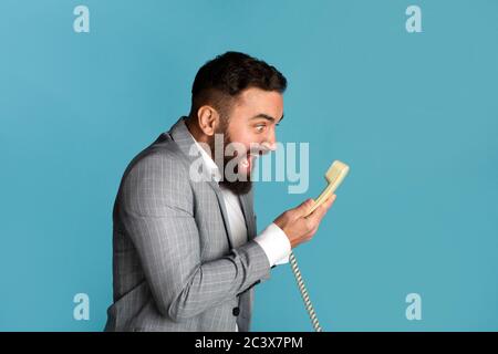 Stress de travail. Un homme d'affaires en colère criait sur un fond bleu dans le récepteur de téléphone fixe Banque D'Images