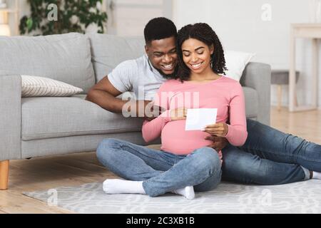 Couple enceinte assis sur le sol et regardant l'échographie Banque D'Images