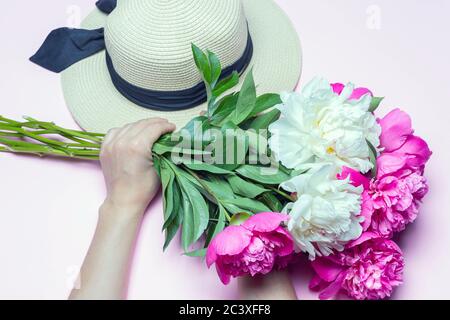 Mélanger le bouquet de pivoines et le chapeau de paille dans les mains femelles isolées sur fond rose. Composition de la pose à plat, gros plan, vue de dessus Banque D'Images