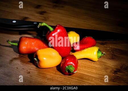 Poivrons doux jaune, rouge et orange vif sur une surface de coupe de bambou recouverte de gouttes d'eau, avec un couteau de chef en arrière-plan. La chute/Autou Banque D'Images