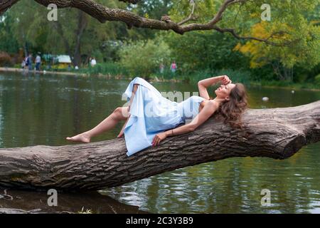 une belle fille aux cheveux bouclés dans une robe bleue est couché sur un grand arbre contre l'arrière-plan d'un Parc avec un lac Banque D'Images