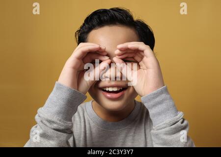 Enfant curieux explorant le monde. Mignon garçon asiatique souriant et regardant à travers les doigts en forme de jumelles et exprimant l'étonnement, contre le dos jaune Banque D'Images