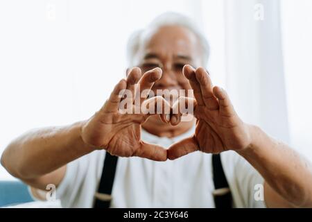 Homme asiatique senior flou souriant et montrant le mouvement de coeur de main pour l'appareil photo dans la pièce confortable à la maison Banque D'Images