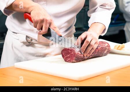 Chef coupant un grand steak de bœuf désossé cru sur une planche à découper blanche. Banque D'Images