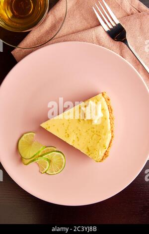 Délicieux morceau de tarte avec les zestes de lime et de tranche de citron vert sur la plaque rose sur table en bois sombre, servi avec une tasse de tisane. Banque D'Images