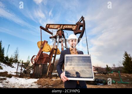 Homme d'affaires portant un manteau de sport, un Jean et un casque blanc, tient un mini module solaire, debout dans un champ de pétrole à côté d'un cric de pompe à huile. Concept de l'industrie pétrolière et source d'énergie alternative Banque D'Images