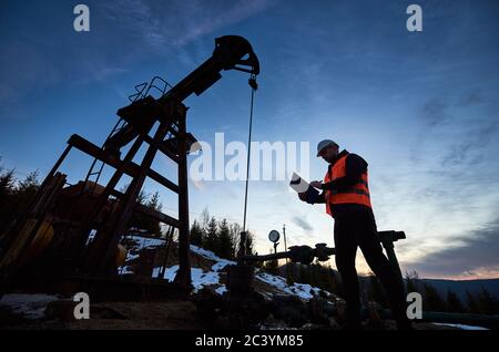 Faible angle de l'ingénieur pétrolier dans le gilet de travail regardant la machine à bascule de pompe à huile et l'écriture sur le presse-papiers. Ouvrier pétrolier vérifiant le travail de la poutre équilibrée de la pompe à pétrole cric sous beau ciel de soirée. Banque D'Images