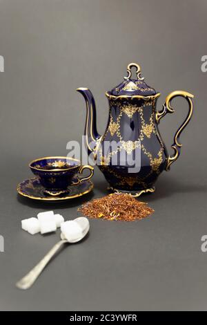 Encore plus de vie avec un magnifique ensemble de thé en porcelaine d'époque bleu cobalt avec motif floral doré, une cuillère avec des cubes de sucre et des feuilles de thé sèches. Banque D'Images