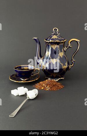 Encore plus de vie avec un magnifique ensemble de thé en porcelaine d'époque bleu cobalt avec motif floral doré, une cuillère avec des cubes de sucre et des feuilles de thé sèches. Banque D'Images