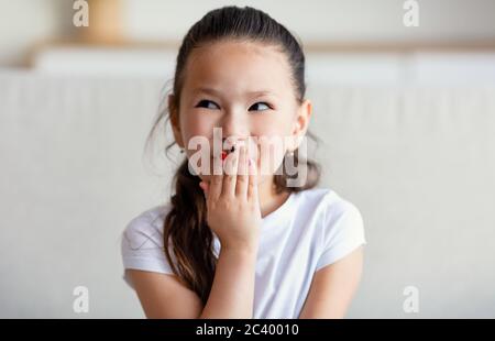 Chinese Girl Chickling couvrant la bouche assis sur un canapé à la maison Banque D'Images