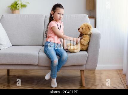 Fille jouant Docteur traiter l'ours jouet assis sur un canapé à l'intérieur Banque D'Images