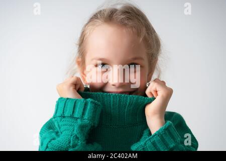 Adorable petite fille cachant la partie inférieure de son visage sous un col épais de pull chaud tricoté. Gros plan studio isolé sur espace de copie blanc Banque D'Images