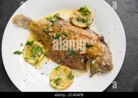 Une semelle de citron entier cuite à la maison, Microstomus kitt, qui a été poêlée dans le beurre avec des tranches de citron, câpres et persil. Présenté sur une plaque blanche Banque D'Images