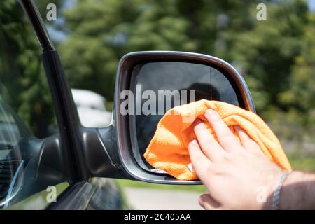 La main du gars essuie le miroir d'aile de voiture avec un chiffon sec. Un chiffon orange nettoie le rétroviseur extérieur. Éponge de nettoyage pour voiture Banque D'Images