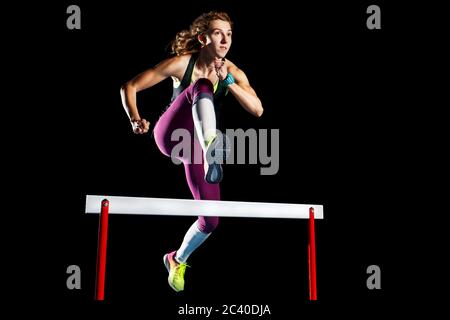 Jeune fille sportive sautant sur l'obstacle isolé sur fond noir. Image de concept de sprint de piste et de champ isolée sur noir Banque D'Images