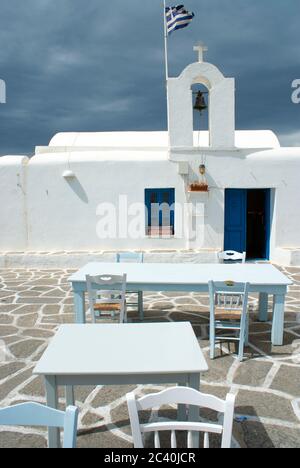 Sur l'île de Paros, des tables et des chaises sont disposées par une taverne sur la terrasse, près d'une ancienne chapelle orthodoxe. Les nuages de tempête se rassemblent. Banque D'Images