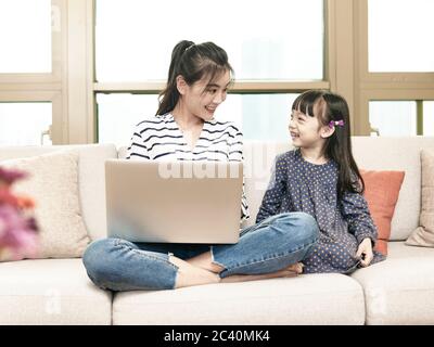 jeune femme d'affaires asiatique travaillant à domicile assise sur un canapé en utilisant un ordinateur portable tout en prenant soin de sa fille Banque D'Images