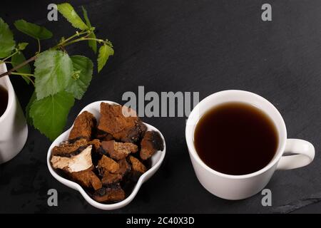 Tasse blanche en céramique de la curatif de chaga aux champignons de bouleau, bol avec morceaux de chaga et feuilles de bouleau sur noir. Café ou thé aux champignons de la chaga. Tendance Banque D'Images