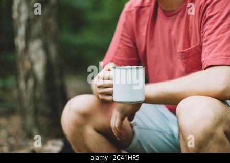 Un homme boit dans une tasse blanche de camping pendant un arrêt dans la forêt. Banque D'Images