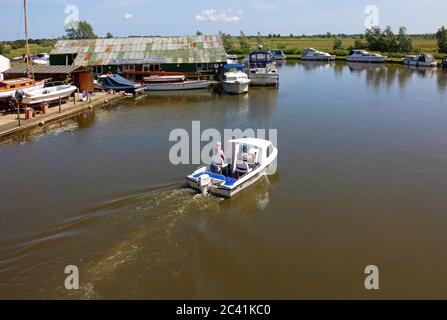 Un couple dans un petit bateau de plaisance faisant la place devant un chantier naval sur la rivière Ant sur les Norfolk Broads à Ludham, Norfolk, Angleterre, Royaume-Uni. Banque D'Images