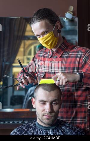 le coiffeur du coronavirus dans un masque protecteur coupe un jeune homme dans un salon de coiffure. Soins capillaires pendant la pandémie du coronavirus, Covid 19, coiffeur Banque D'Images