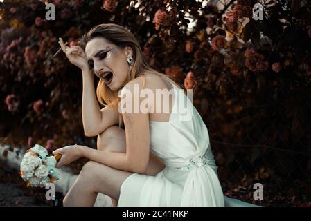 la mariée en robe blanche pleure, tenant une cigarette dans ses mains, tenant une bouteille d'alcool. Sur fond floral de Banque D'Images