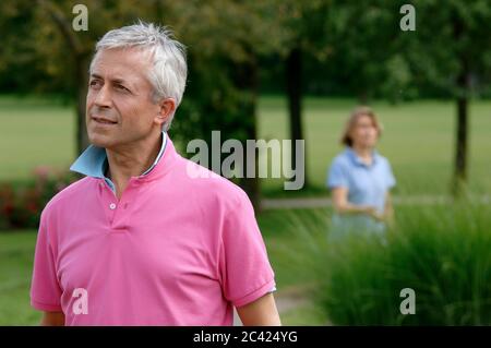 Homme aux cheveux gris dans un polo rose regarde dans la distance, en arrière-plan une femme - loisir - détente Banque D'Images