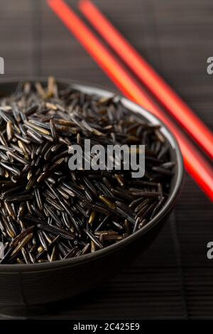 Tas de grains de riz sauvage non cuits, crus, noirs dans un bol noir avec des baguettes rouges sur fond noir de natte de bambou, foyer sélectif Banque D'Images