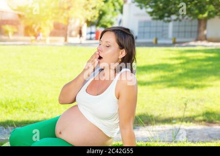 Femme enceinte bâillements. Belle future mère prépare pour la naissance de bébé, fatigué et essayant de se détendre, assis sur une herbe verte dans un parc ensoleillé en plein air Banque D'Images