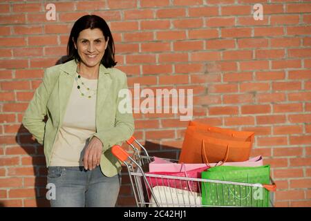 Une femme âgée se tient devant un mur de briques avec un panier rempli de sacs d'achats Banque D'Images