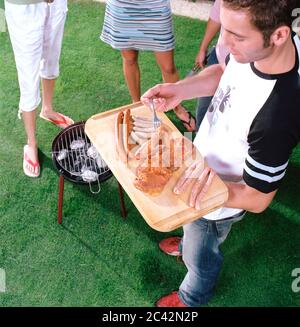 Jeune homme avec de la viande et des saucisses sur bois - jeunes à côté du grill dans le jardin - barbecue Banque D'Images