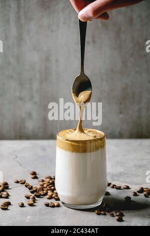 Dalgona café mousseux tendance coréenne boisson latte de lait avec mousse de café dans tasse de verre, mousse qui coule de la cuillère. Table de textures grises. Copier l'espace Banque D'Images