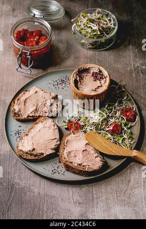 Sandwiches et bol en céramique de pâté de foie de poulet maison avec couteau en bois, tomates séchées et salade verte sur assiette grise sur texture sombre Banque D'Images