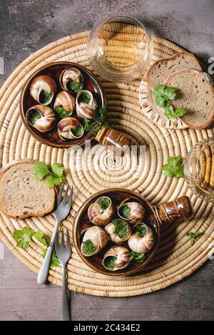 Escargots de Bourgogne - escargots au beurre d'herbes, plat gastronomique, dans des casseroles traditionnelles en céramique avec coriandre, pain, verre de vin blanc sur la paille napki Banque D'Images