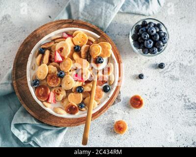 Plats tendance - céréales à crêpes. Lait et crêpes dans le bol. Petites crêpes aux céréales avec baies dans une assiette de métier sur fond de ciment gris. Copier sp Banque D'Images