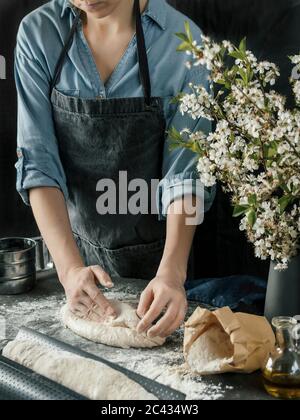 Jeune femme en chemise bleue et tablier gris faisant de la pâte à pain. Les mains des femmes moulant de baguette sur la cuisine sombre. Table noire avec ingrédients alimentaires - flo Banque D'Images