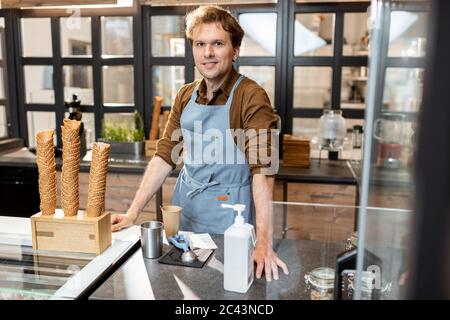 Portrait d'un vendeur ou d'un propriétaire d'entreprise gai vêtu d'un tablier debout au comptoir d'une pâtisserie ou d'un café Banque D'Images