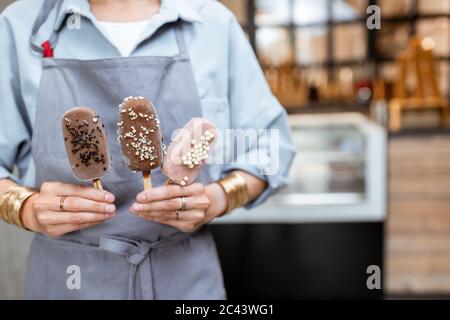 Femme vendeur tenant de la crème glacée au chocolat sur un bâton dans la boutique, gros plan sur les mains et la crème glacée Banque D'Images