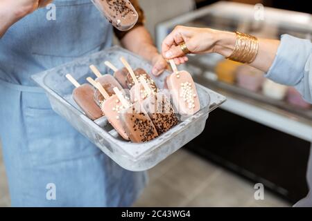 Vendeur tenant un plateau avec de la glace au chocolat sur un bâton dans la boutique, gros plan sur de la glace sur un fond bleu de tablier Banque D'Images