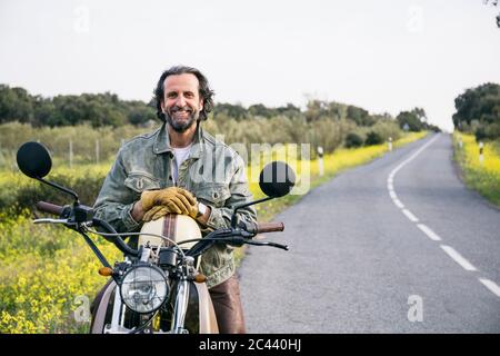 Homme barbu souriant assis à moto à la campagne Banque D'Images