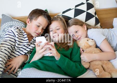 Photo de groupe de trois enfants qui se trouvent ensemble sur le lit et qui regardent le téléphone portable Banque D'Images