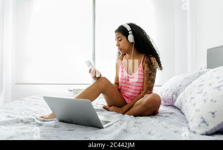 Belle jeune femme avec casque, smartphone et ordinateur portable dans le lit Banque D'Images