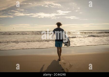 Costa Rica, province de Puntarenas, Puntarenas, vue arrière de surfeur mâle marchant vers l'océan avec planche de surf en main Banque D'Images