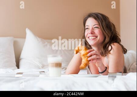 Bonne jeune femme tenant un croissant tout en prenant le petit déjeuner au lit Banque D'Images