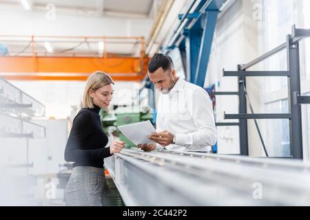 Homme d'affaires et femme avec des papiers examinant des tiges de métal dans le hall d'usine Banque D'Images